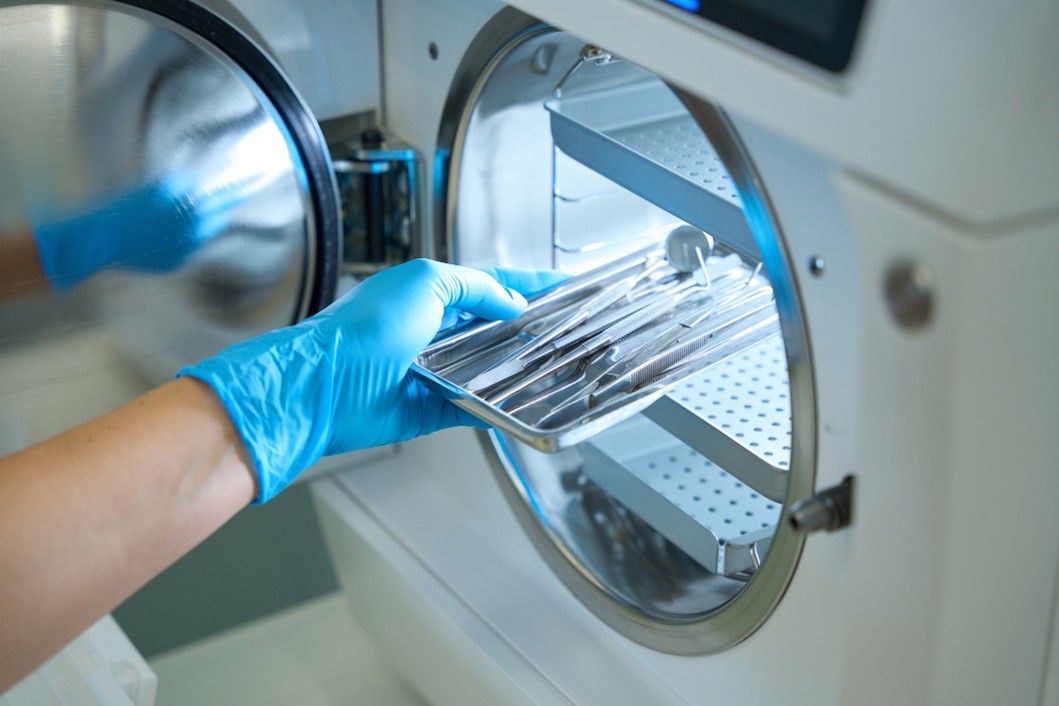 A gloved hand is placing a tray of dental instruments into a sterilization machine.